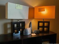 Two table lamps with black geometric bases and rectangular fabric shades (one white, one orange) alongside three gray and white striped cat figurines on a dark surface