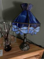 Photo showing Tiffany style stained glass lamp with blue floral shade, reed diffuser in dark brown bottle, and three clear glass vases on wooden surface.