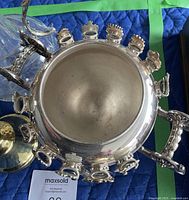 Top view of silver-plated spooner showing round body with multiple enamel crest badges attached around it and attached spoons hanging in front.