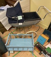 Photo showing vintage metal baby pram with blue fabric canopy and large wheels alongside blue wooden doll cradle and various books in background.