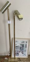 Two brass floor lamps leaning against a wall beside a framed garden print resting on the floor.