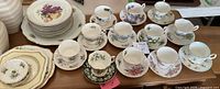 Set of fine bone china teacups and matching saucers with various floral designs, arranged in front of stacks of plates and a large fruit-design serving platter.