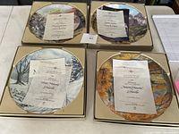 Four collectible plates in original boxes, each with a certificate of authenticity placed on top, showing titles and artists: Autumn Memories, Winter Memories, The Grist Mill, and Majestic Rockies.