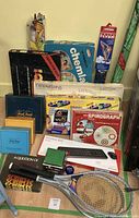 Photo showing various vintage games and toys, including Trivial Pursuit, ChemLab Explorations, Lite Brite, Spirograph, Scruples, and A Question Of Trivia. Two wooden tennis rackets are visible along with a boxed Wireless 800 keyboard and a packaged Flyer kite.
