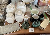 Overview of kitchen items on table including ceramic canisters, pottery creamer and sugar dish, glass pitcher, mugs, butter dish, and trays.
