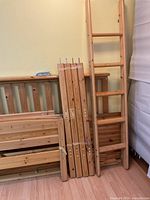 Disassembled parts of wooden loft bed showing ladder, side rails, and other components stacked against the wall.