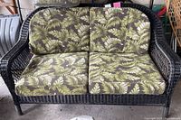 Full view of brown wicker loveseat with green floral cushions on seat and back, showing overall condition.