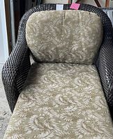 Front-facing view of the dark brown wicker lounger with green cushioned pad with leaf pattern, showing the full seat and armrests.