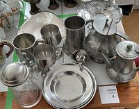 Top view of the 13-piece pewter and metalware set arranged on a wooden table showing teapots, cups, plate, pitcher, glass decanter, metal bowl and tray.