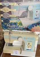 White Multi-Tasker sewing machine with box in background showing product features like built-in stitches, buttonhole making, included presser feet, and multiple stitch functions.