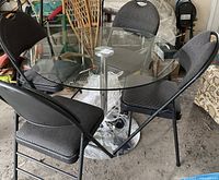 Photo showing round glass top table with chrome base and four black folding chairs around it in a cluttered storage area.