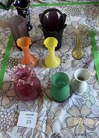 Nine colorful glass vases arranged on a floral-patterned tablecloth, showing various shapes and colors