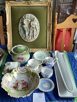 Full lot arranged on table showing framed 3D porcelain wall art and various porcelain cups, bowls, plates, and serving pieces.