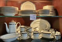 Full shelf view showing stacked dinner and salad plates on top shelf, cups and saucers, teapot and serving platter on bottom shelf