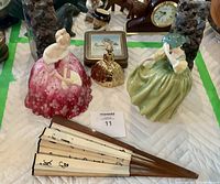 Two Royal Doulton ceramic figurines, a brass hand bell shaped like a lady, two large amethyst crystals, and a hand fan with Asian script laid out on a padded surface.
