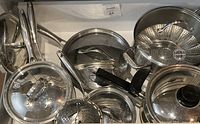 Overhead photo showing all five cooking pots each with a lid, two perforated steamer trays, and the double boiler pot with lid inside a white drawer.