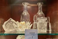Front view of two Waterford crystal whiskey decanters, ice bucket with tongs, crystal votive holder, and frosted ice cube tray on glass shelf inside a wooden cabinet.