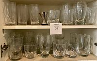 View of two shelves in a cabinet containing multiple clear drinking glasses and tumblers in various styles and sizes with a metal jigger in the center top shelf.