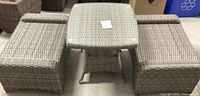Square wicker outdoor table with glass top flanked by two matching wicker seats, light brown color and woven pattern.