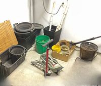 Photo of various black plastic planters, green bucket, hand air pump, small pots and white birch branch stakes on a concrete floor in a storage area.