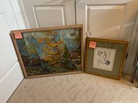 Two framed art prints leaning against a white door on carpeted floor, one titled 'Laurentian Woods' with autumn landscape in a wood frame and the other a botanical print in a gold frame with green mat