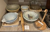 Overall view of glass kitchenware arranged on countertop including differing sized Pyrex bowls, pie plates, baking dishes, a wooden rolling pin, and pastry cutter.