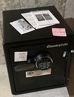 Front-facing view of black SentrySafe with digital keypad, handle, maxsold label 167, dimensions tag '16 x 16 x 12', manual and hardware pack on top