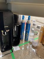 Two black SodaStream Tera soda makers alongside two new CO2 cylinders and six transparent 1L Fuse bottles with white caps and bases on a countertop.