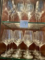 25 large clear glass wine glasses displayed on two glass shelves inside a cabinet.