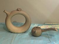 Front view of vintage clay wine decanter with circular center and cork lid next to clay teapot with wooden handle on blue surface