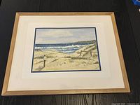 Framed watercolour beach scene print by Cornell, showing sandy beach with grassy dunes and ocean waves, light wood frame and cream matting.