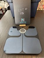 Top angled view of black Fitbit scale with a box labeled 'The Next Step Beyond' in background and lot number written on box