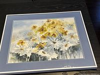 Watercolour print showing yellow and white flowers with green stems framed with a blue matte and silver frame laying flat on a dark surface.