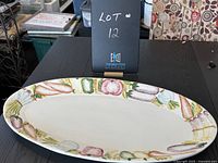 Top-down view of large oval serving plate with colorful vegetable motif around the rim, placed on black table with a lot number sign behind it.