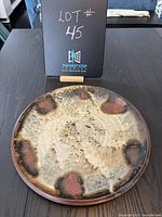 Top-down photo of round ceramic plate with textured glaze and brown spots on a dark wood surface, labeled lot number 45 behind it.