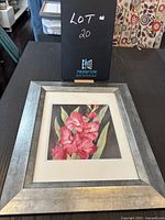Framed watercolour painting showing pink gladiolus flowers titled "Firefly" by Margaret Gee, displayed on a dark table background with patterned fabric.