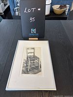 Full view of framed still life print featuring wooden chair on patterned rug, with lot number sign in background.