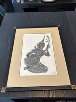Framed Thai rubbing artwork featuring a traditional musician kneeling and playing an instrument, shown on a table surface.