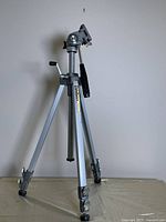 Full view of the Velbon VG-3 tripod showing three silver metal legs with locks and black rubber feet. The tripod head has a black handle for adjusting the angle.