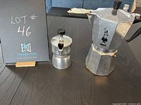 Photo showing both espresso pots side by side on a black table, with lot number signage in background.