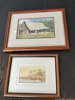 Two framed watercolour prints leaning on a surface. Larger print shows a barn-like building and smaller shows a landscape.