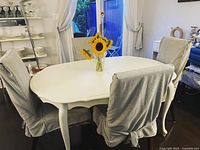White oval dining table with scalloped edges and queen anne style legs, surrounded by slipcovered chairs, in a room with shelves of plates.