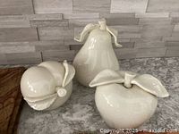 Three white ceramic decorative fruits (pear, apple, peach) arranged on countertop with gray tiled backsplash.