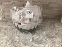 Large clear glass punch bowl filled with fabric and eight small glass cups placed in front on a counter.