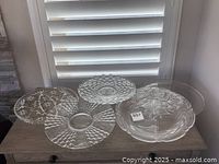 Five clear glass platters with various patterns placed on a wooden surface near a window with closed blinds, showing varying designs and sizes.