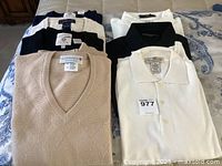 Photo shows seven medium-sized men's V-neck sweaters and polo shirts neatly folded on a bed. Includes a beige V-neck crew neck sweater in front of six folded polo shirts in various colors - striped navy/white, navy, black, and white.