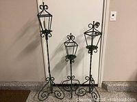Three green metal standing lanterns of different heights arranged against a wall, showing their full stands and candle holder frames.
