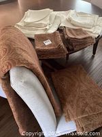 Photo showing brown paisley tablecloth and matching napkins stacked on a white chair and wooden table.