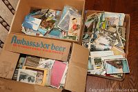 Three cardboard boxes, including one labeled “Ambassador beer,” filled with loose postcards.