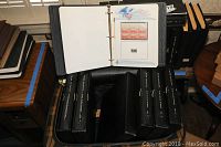 Open Pullman suitcase showing six black stamp collecting binders and one smaller black binder inside, arranged on a wooden surface.